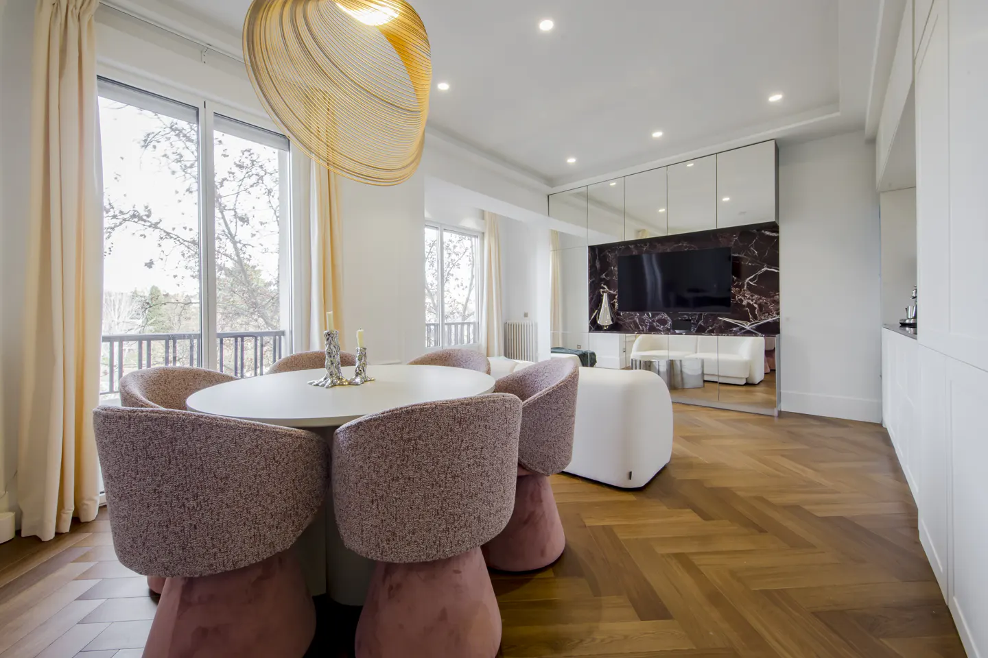 Bright, modern dining room with pink chairs around a white table, wood floors, and a mirrored wall reflecting a living area.