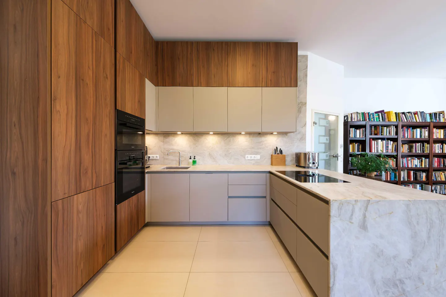 Modern kitchen with wood and beige cabinets, marble countertops, and a large island. A bookcase is visible in the background.