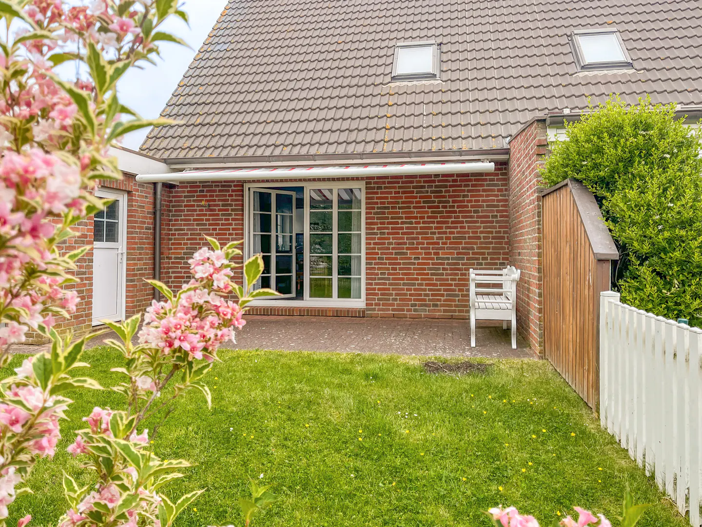 Backyard view of a brick house with a white bench, awning, and open sliding glass door. Pink flowers are in the foreground.