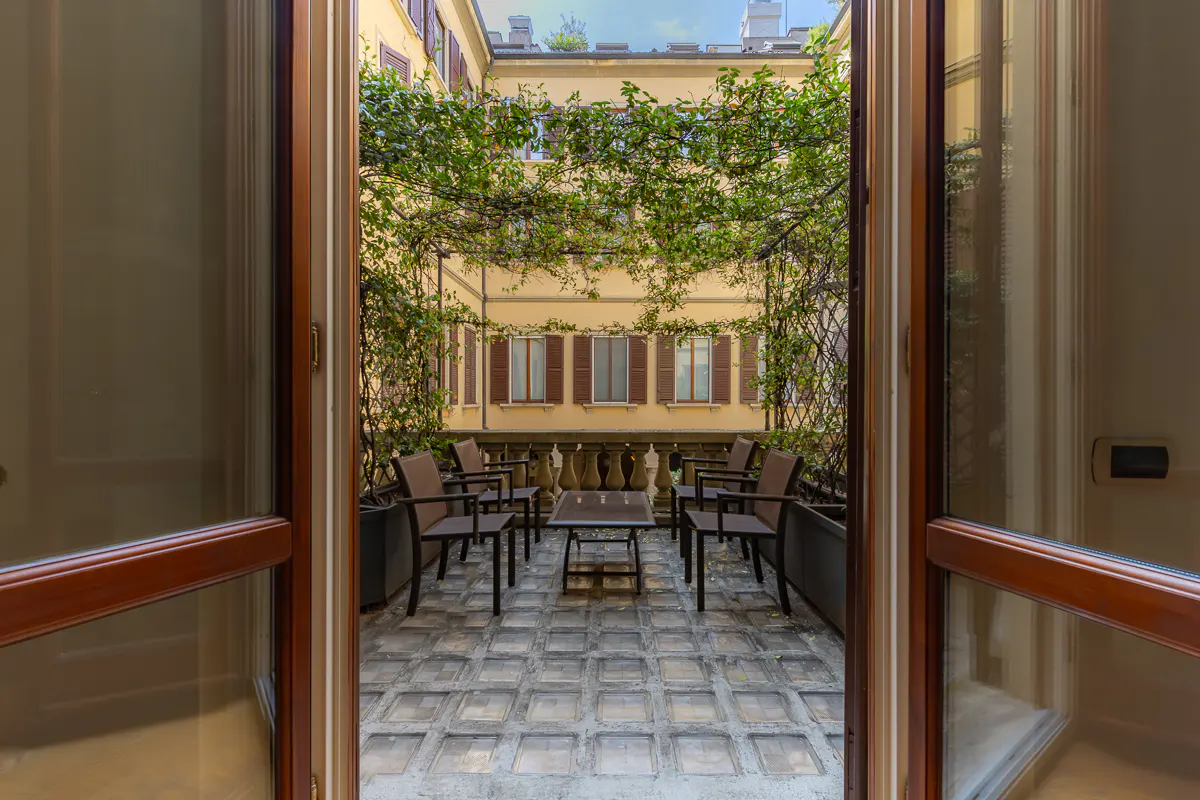 View through open doors to a courtyard with chairs, table, and climbing vines on a yellow building.