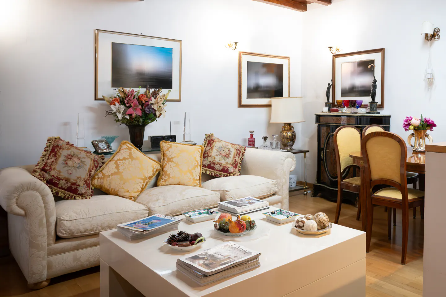 Living room with a white sofa, decorative pillows, and a white coffee table with magazines and snacks. Art hangs on the white walls.