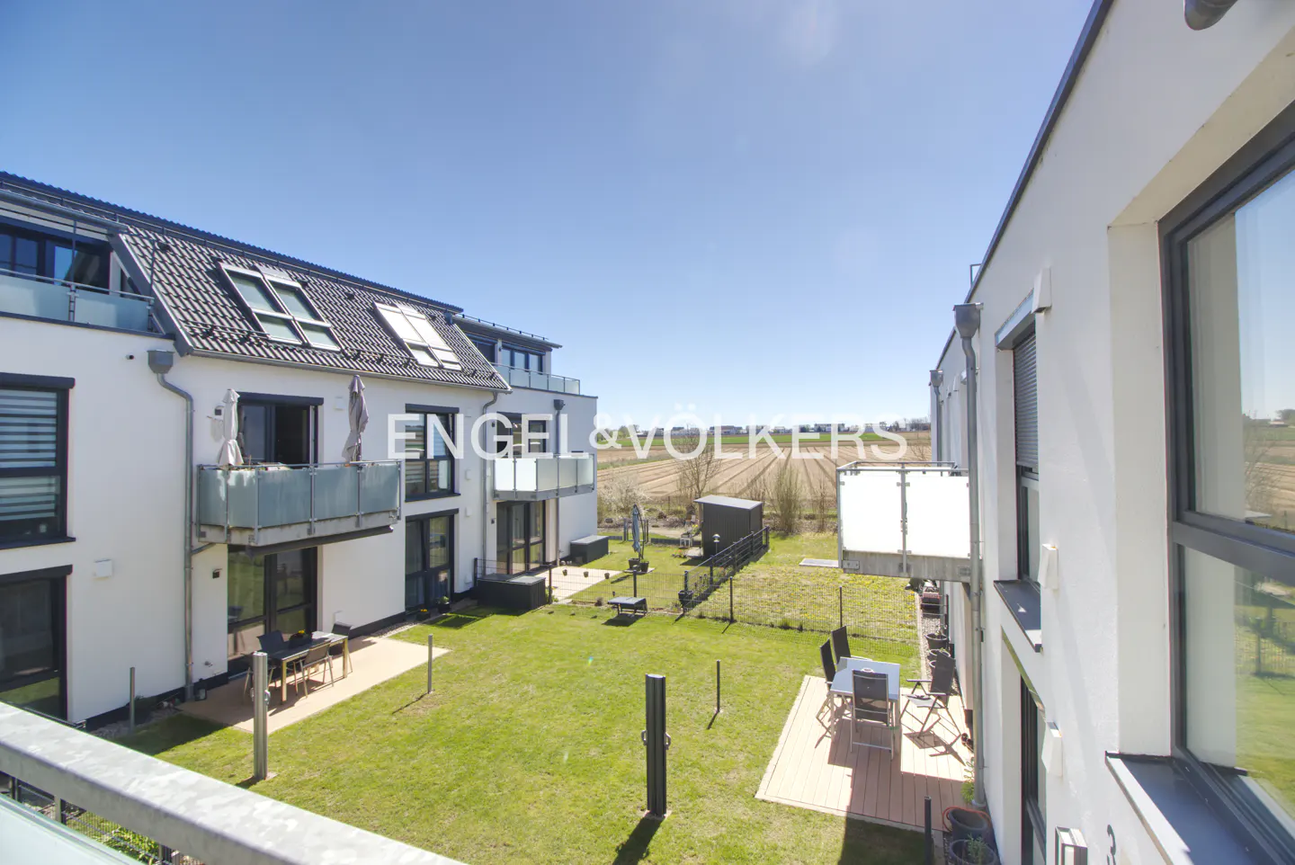 View of modern white townhouses with balconies overlooking a green lawn and patio furniture under a clear blue sky.