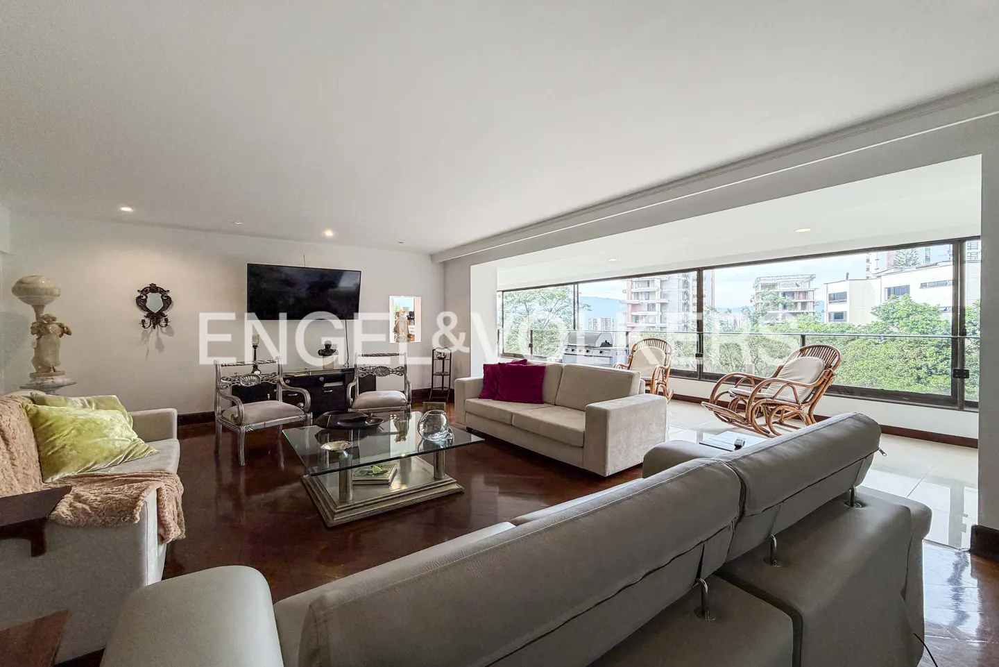 Bright living room with sofas, glass table, TV, and floor-to-ceiling windows overlooking trees and buildings.