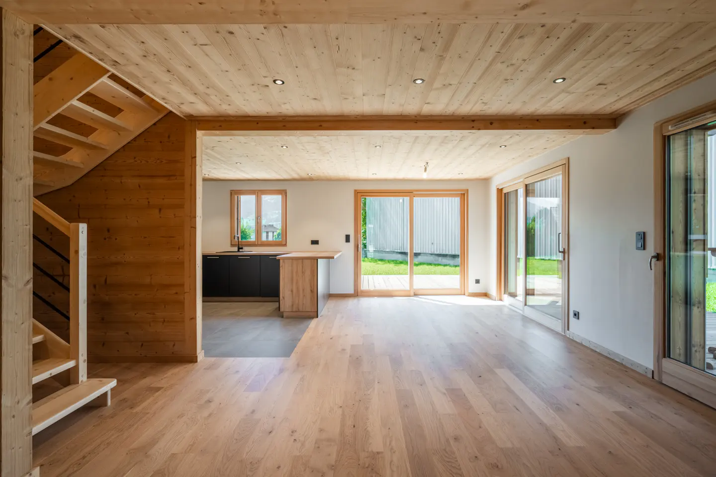 A bright, modern interior with light wood floors, walls, and ceiling. A staircase is on the left, and sliding glass doors lead to the outside.