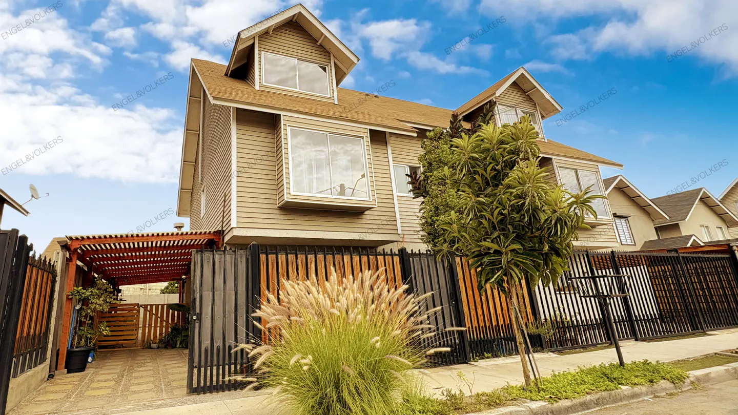 A two-story beige house with a brown roof, black fence, and green tree under a blue sky.