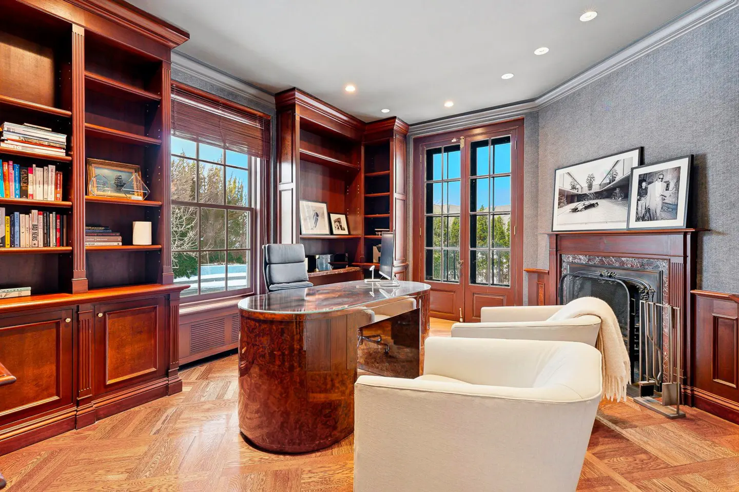 A home office with wood floors, built-in bookshelves, a desk, and two white chairs. A fireplace is visible in the background.