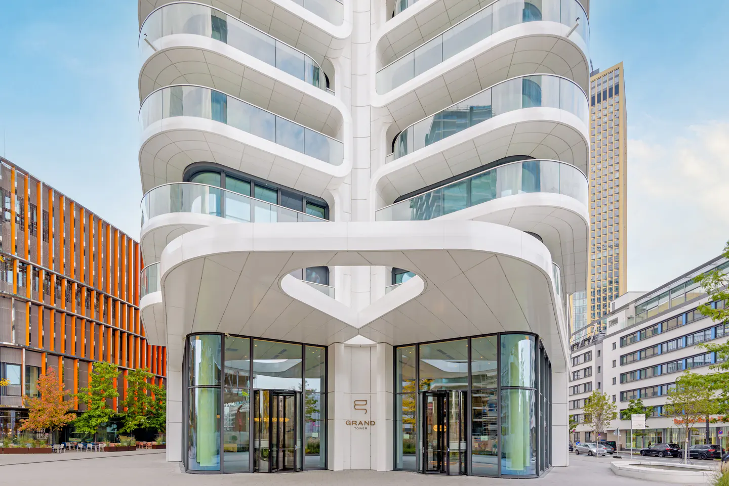 Modern white apartment building with curved balconies and glass doors at the entrance. "Grand Tower" logo visible.