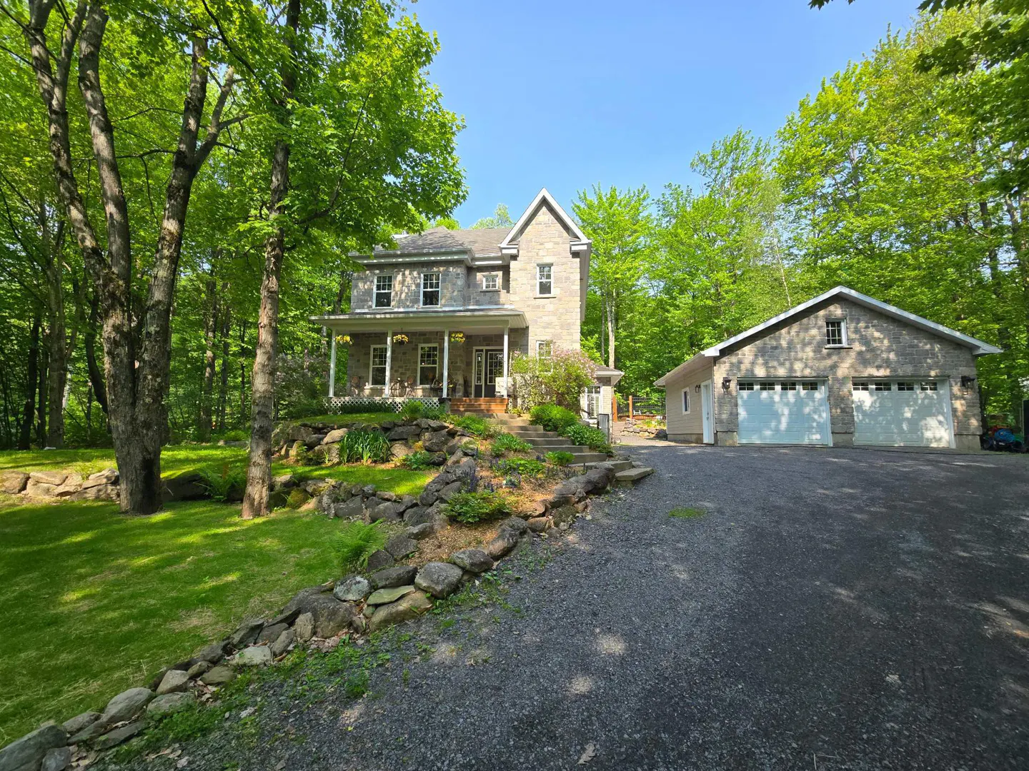 Exterior view of a two-story stone house with a porch, surrounded by green trees and a stone wall. A two-car garage is on the right.
