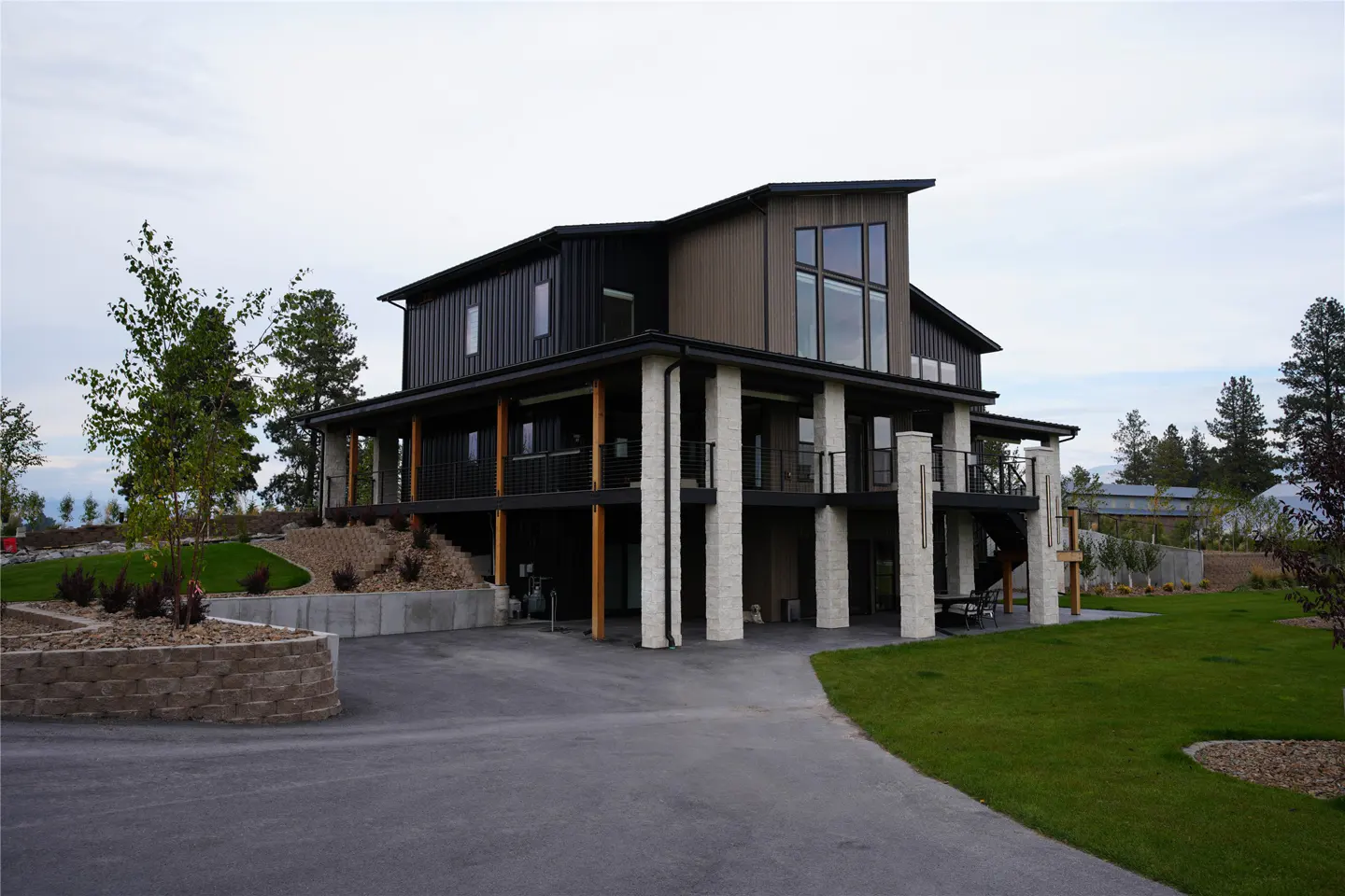 Two-story modern house with black siding, light brown accents, and large windows, supported by white pillars. A driveway leads to the house.