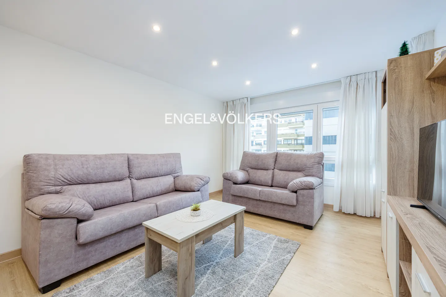 Living room with two grey fabric sofas, a light wood coffee table, a grey patterned rug, and white curtains.