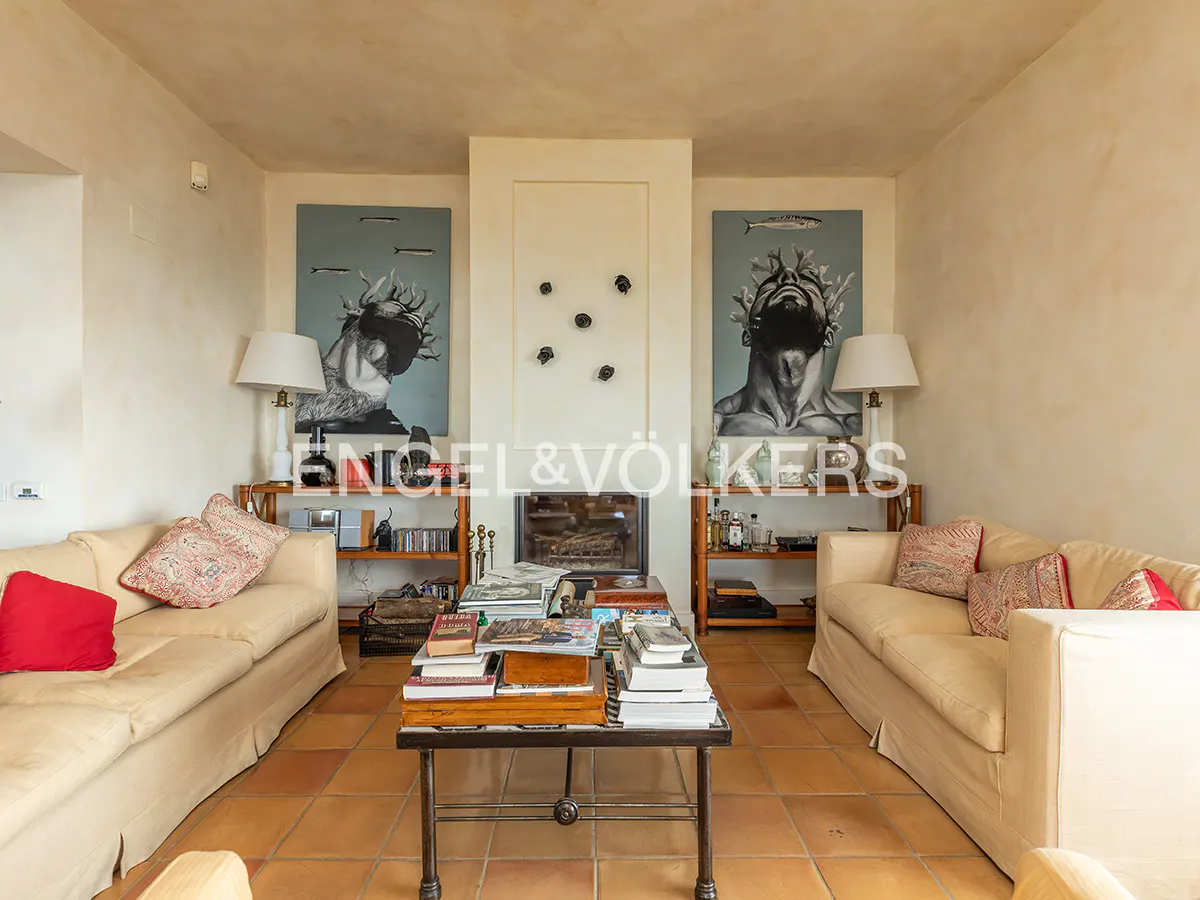 Living room with beige sofas, a fireplace, and artwork. A coffee table is piled with books and magazines. The floor is tiled in terracotta.