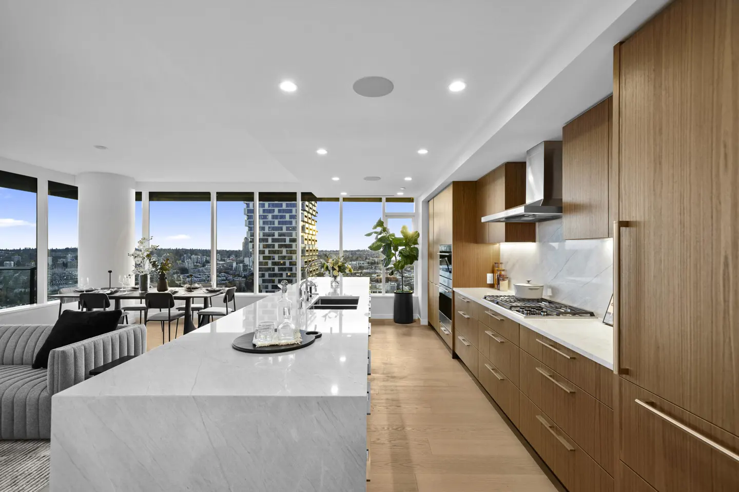 Bright, modern kitchen with wood cabinets, marble island, and city views from large windows. Dining table and gray sofa visible.