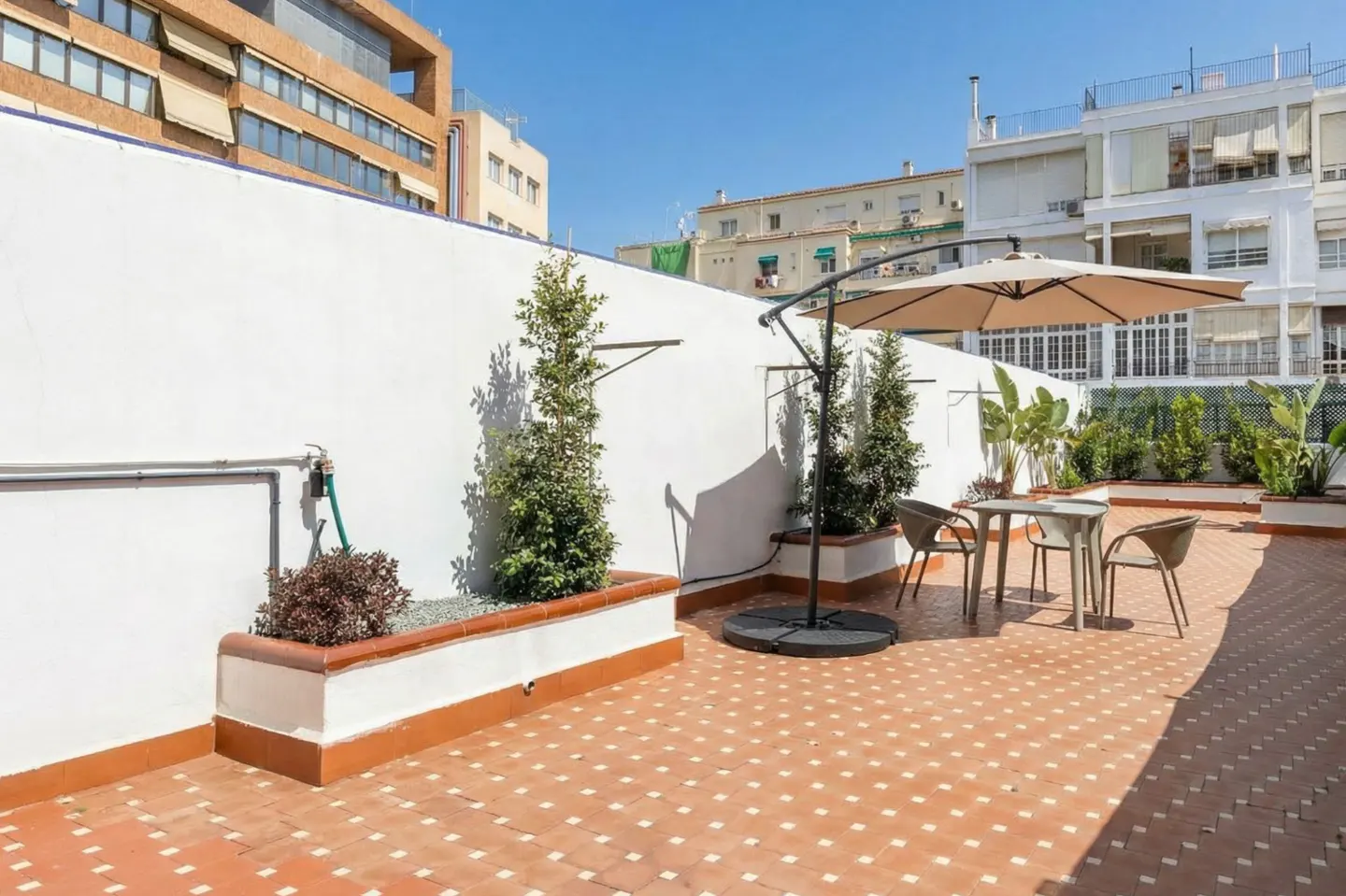 Outdoor patio with red tile flooring, white walls, and potted plants. A table and chairs sit under a beige umbrella. Buildings are visible in the background.
