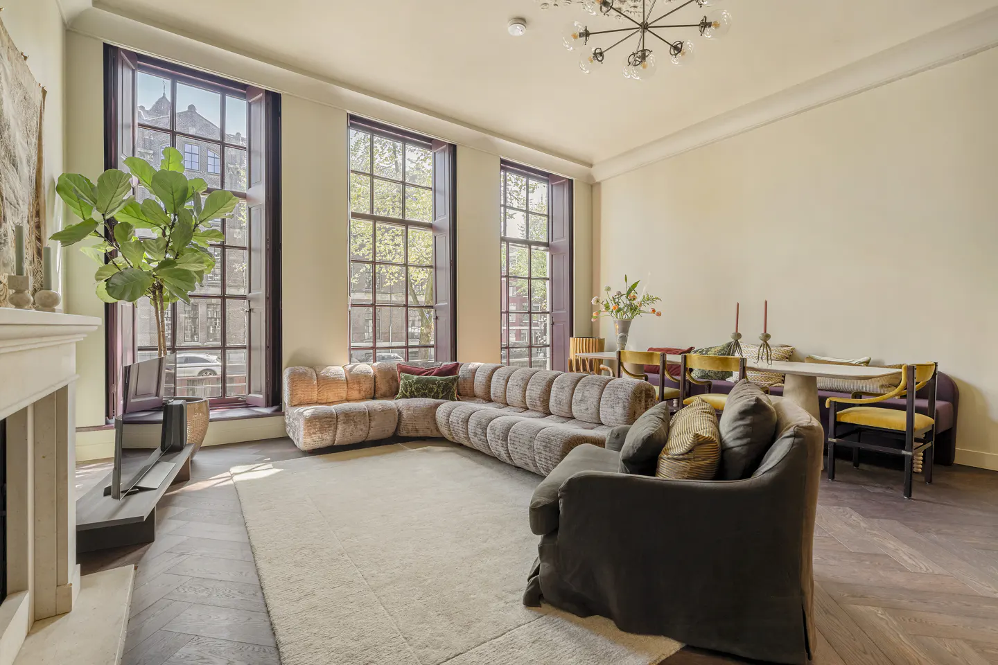 Bright living room with large windows, a beige sectional sofa, and a dark armchair. A dining table with chairs is in the background.