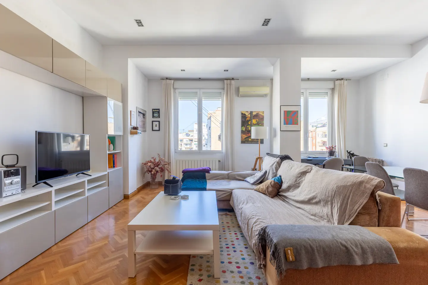 Bright living room with a large sectional sofa, white walls, and herringbone wood floors. A white media console and coffee table add to the modern aesthetic.
