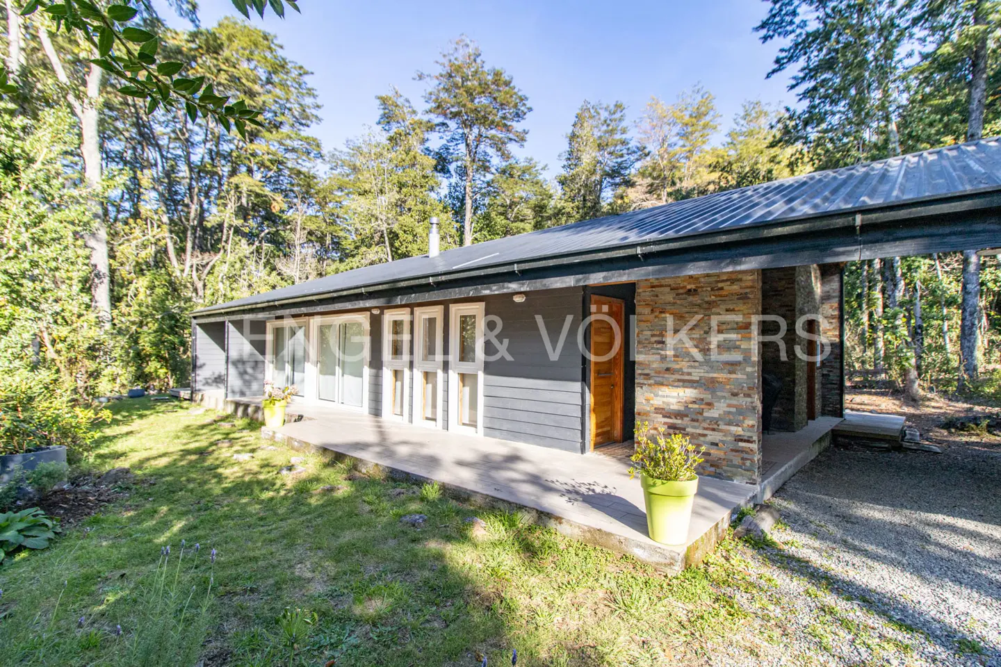 Exterior view of a gray house with a stone accent wall, a wooden porch, and a green lawn surrounded by trees.