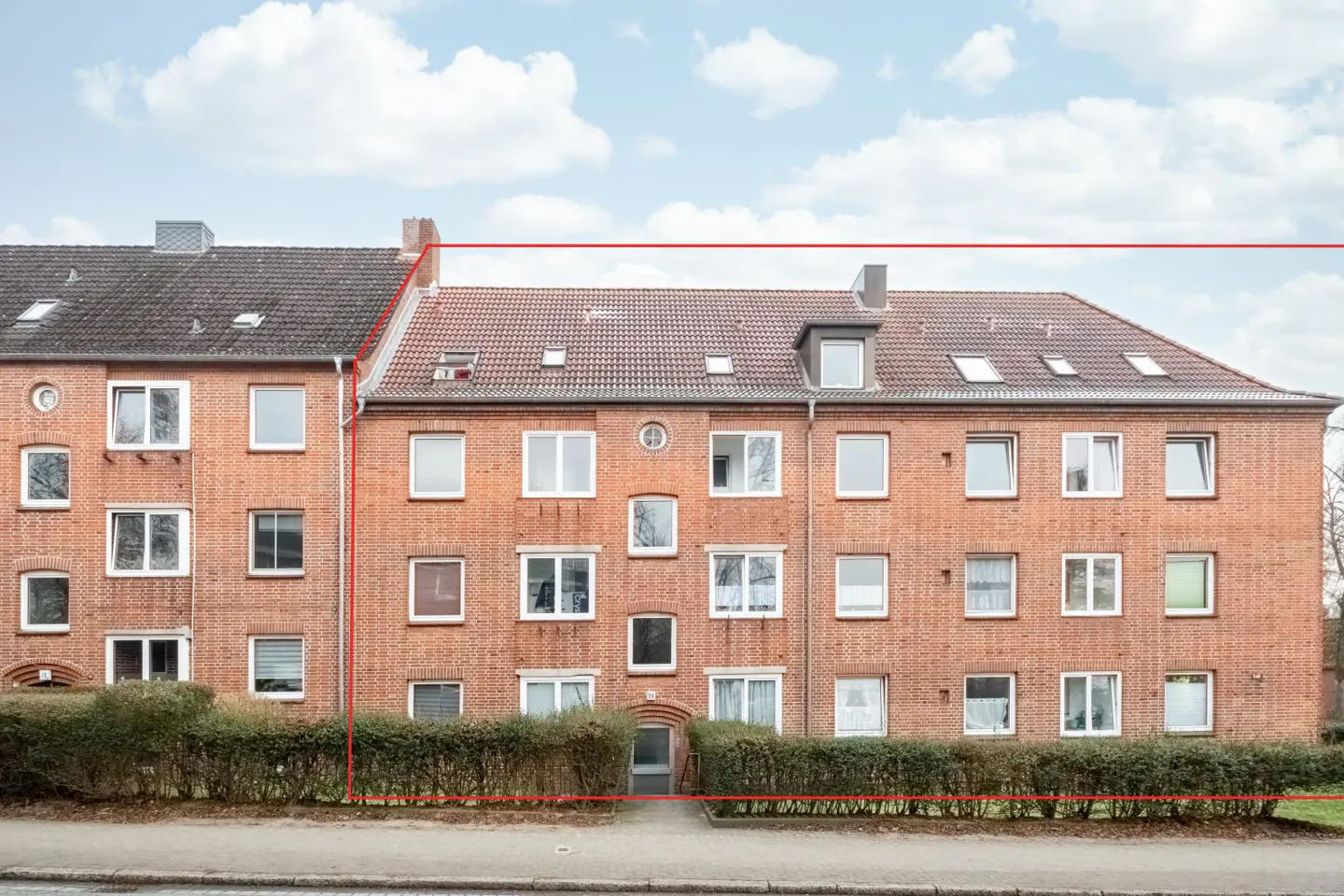 Exterior view of a three-story brick apartment building with a red tile roof and white-framed windows under a cloudy sky.
