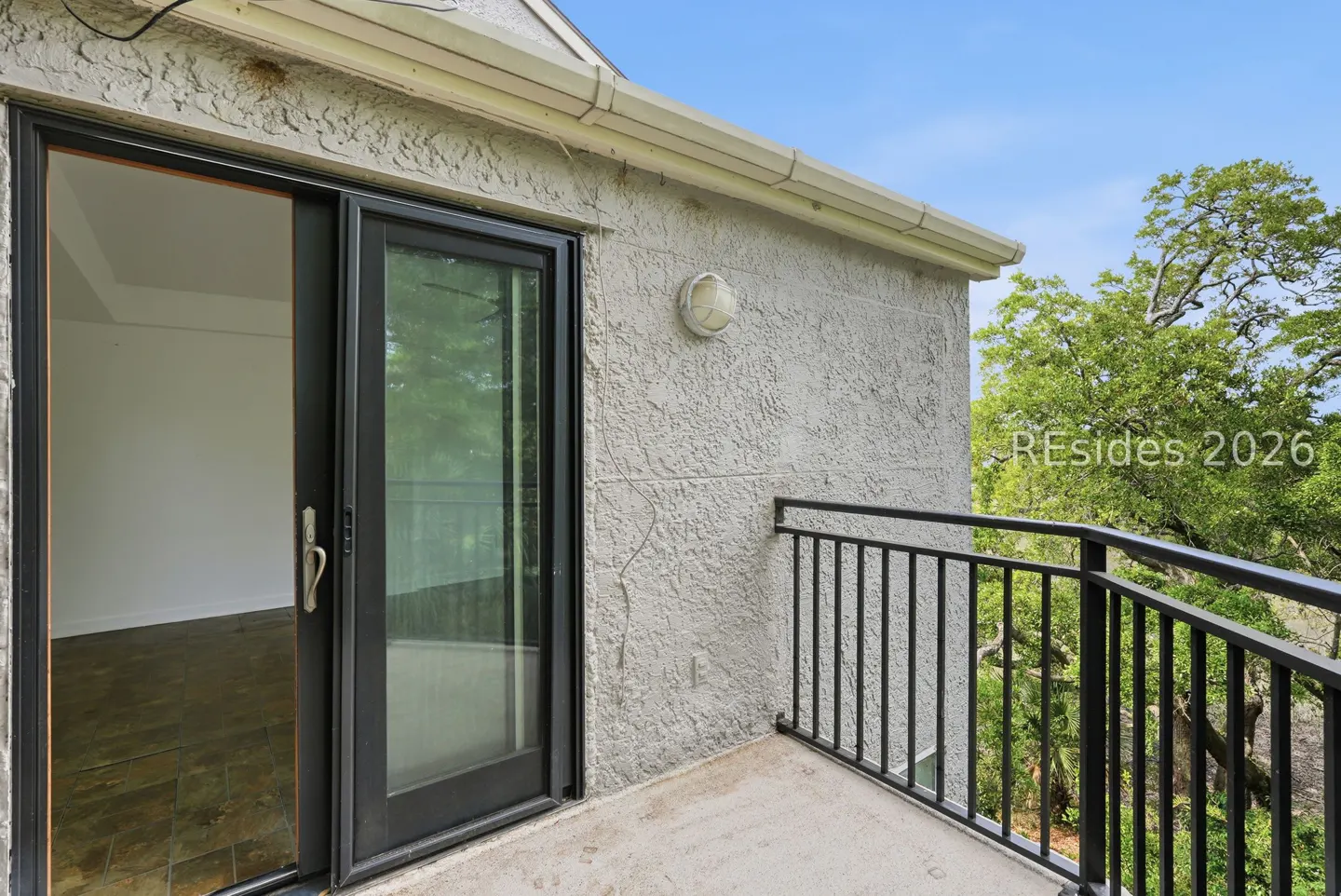 Balcony with black metal railing, sliding glass door open to a room with tile floor. Trees and blue sky in the background.