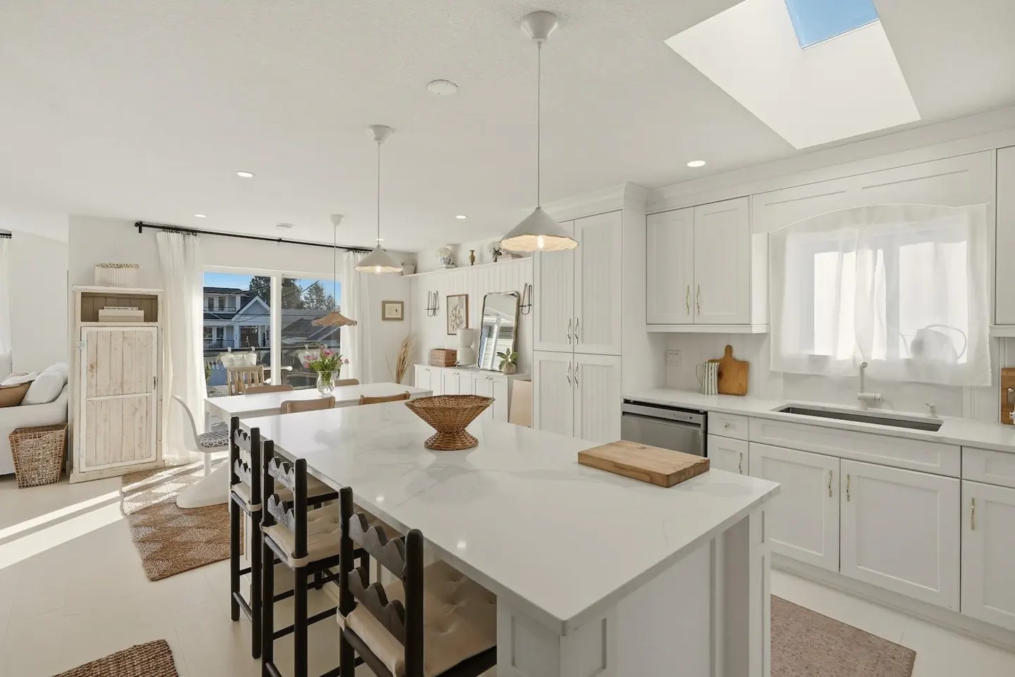 Bright, white kitchen with island, black chairs, and skylight. Cabinets line the wall, and a dining area is visible through a window.