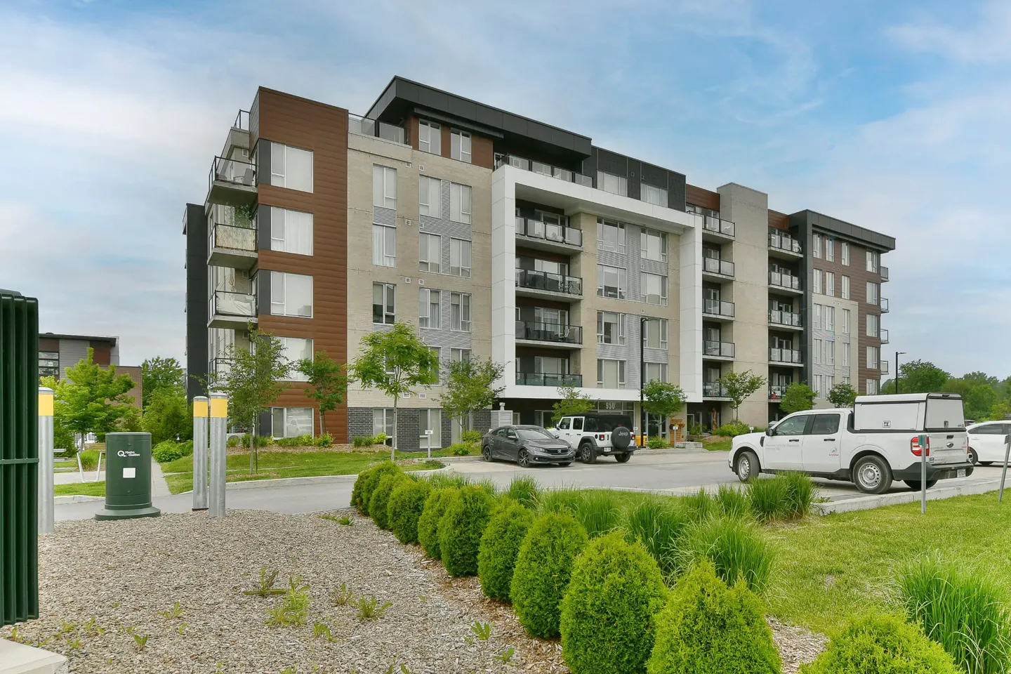 Modern multi-story apartment building with brown and grey facade, balconies, and a parking lot with cars in front.