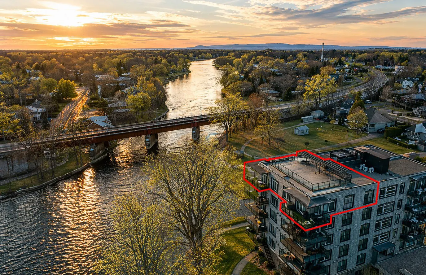 Aerial view of a modern condo building with a red outline, near a river and bridge at sunset.