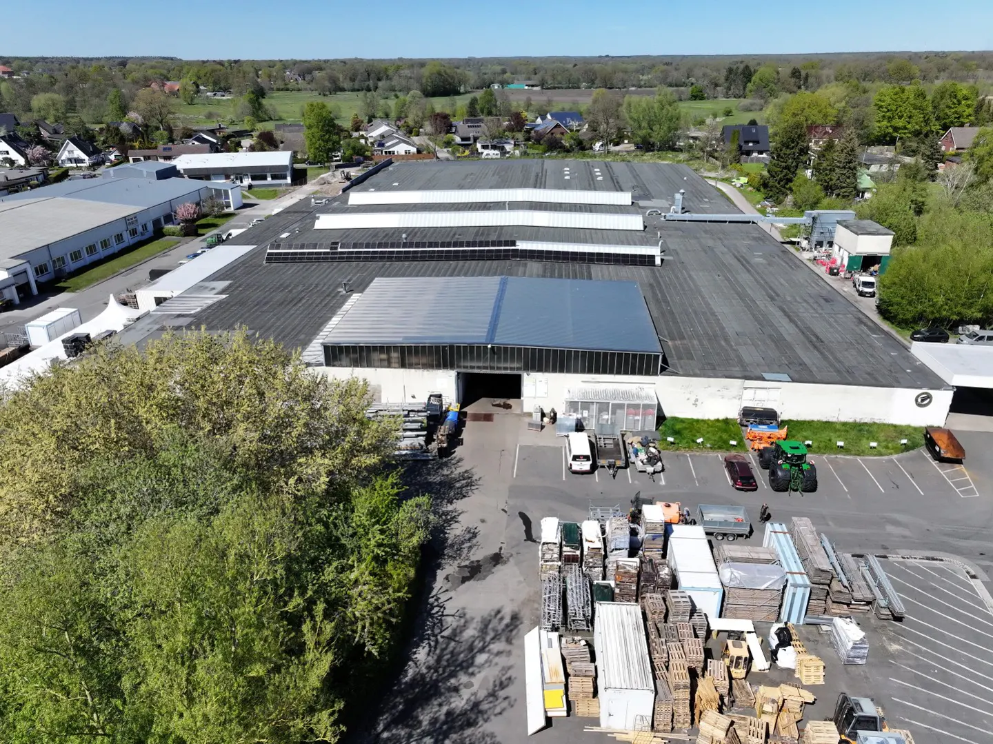 Aerial view of a large, gray-roofed industrial building with skylights, surrounded by trees and a parking lot with vehicles and stacked materials.
