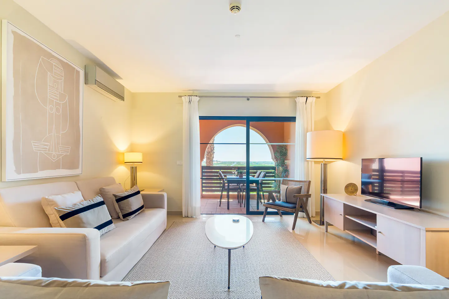 Bright living room with beige sofa, striped pillows, and a glass door to a balcony with outdoor seating. Modern art and a TV add to the room's appeal.