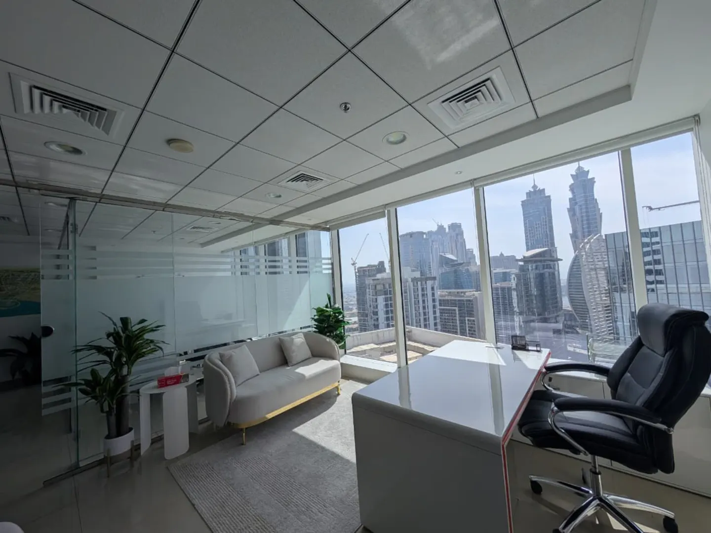 Bright office space with a white desk, black chair, and a grey sofa near a large window overlooking a city skyline.