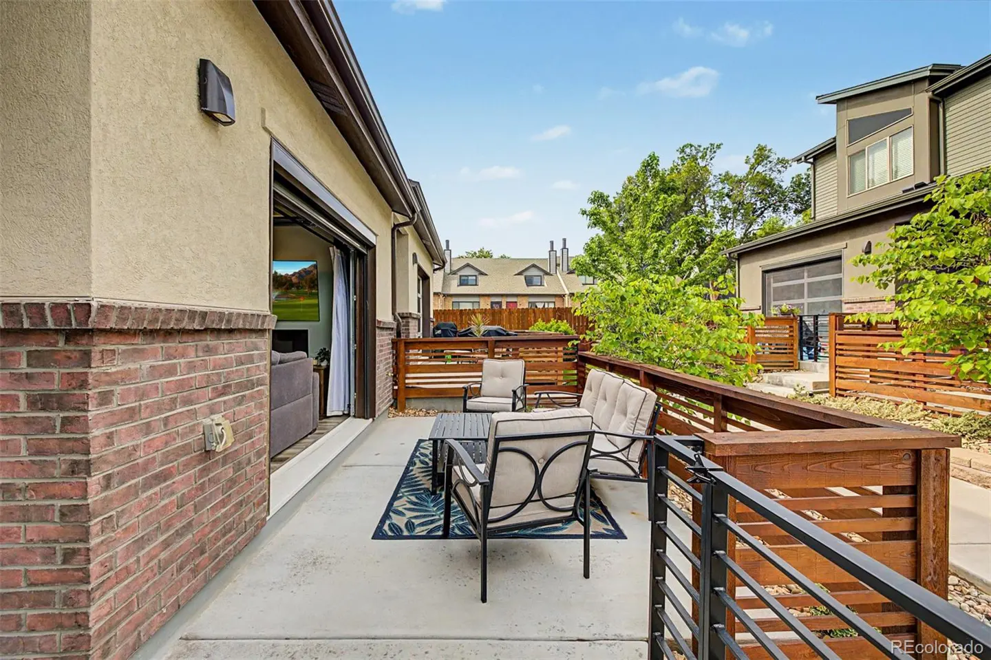 Outdoor patio with seating area, including a sofa, chairs, and a table on a blue rug. A sliding glass door leads to the interior. Wooden fence surrounds the patio.