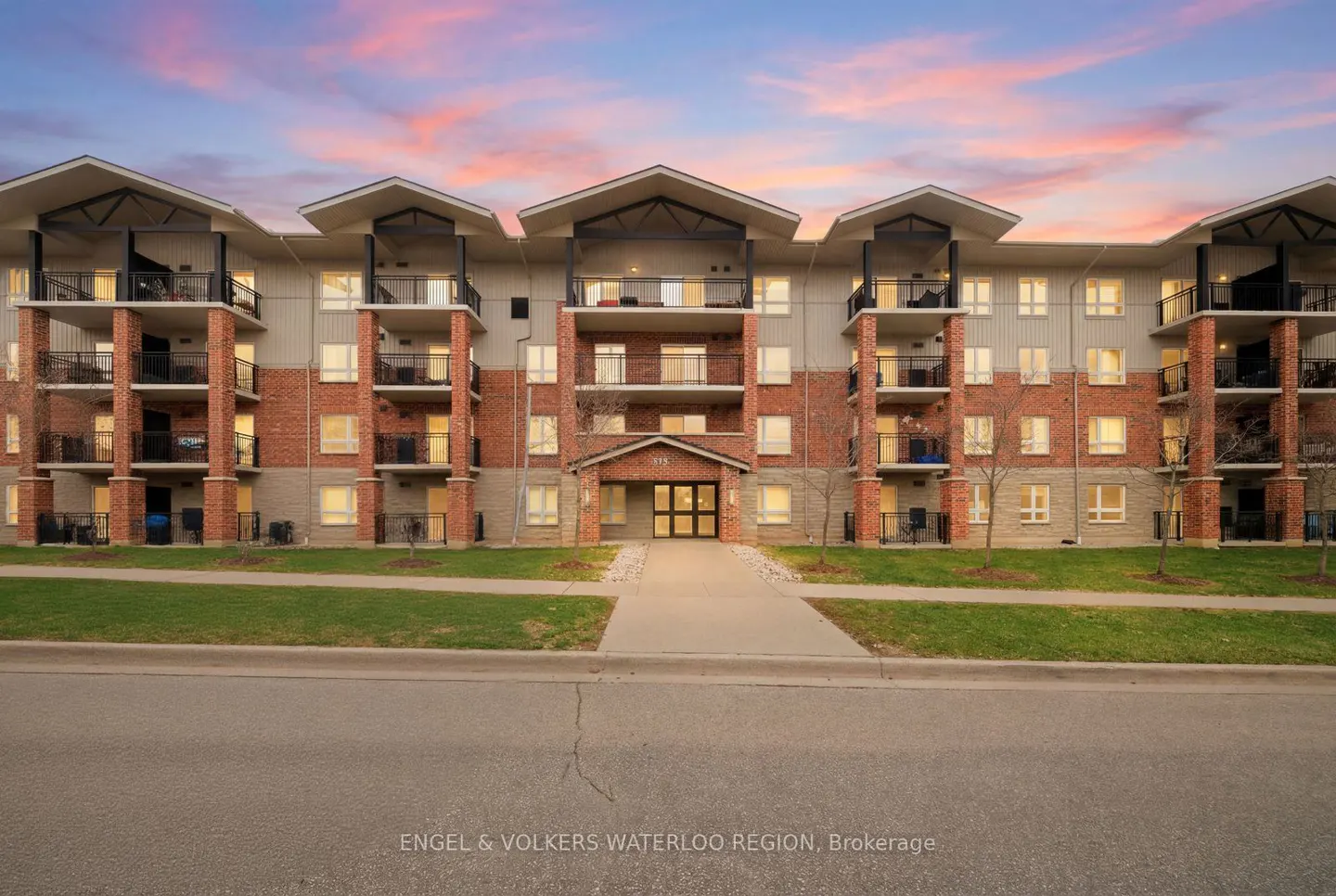 Three-story apartment building with brick and gray siding, black balconies, and a sidewalk leading to the entrance. The sky is pink and orange.
