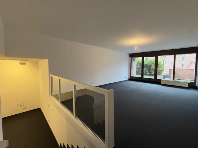 Interior view of a modern home with black carpet, white walls, and large windows overlooking a green yard. A glass railing overlooks the living space.
