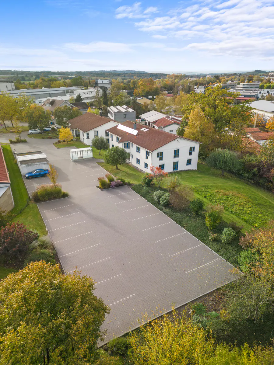 Aerial view of a white building with a red roof, a large parking lot, and green trees under a blue sky.