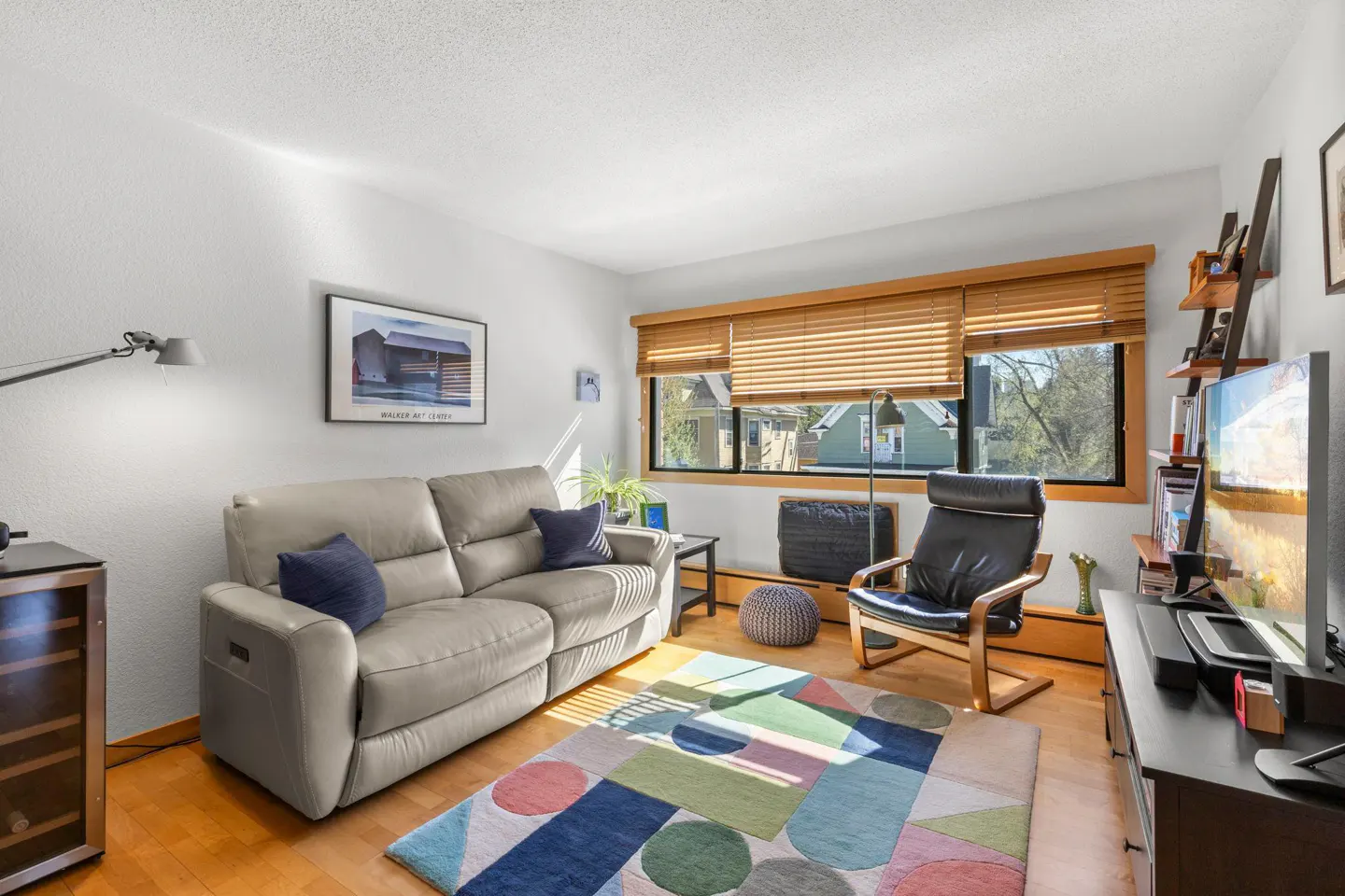 Living room with a gray sofa, blue pillows, and a colorful geometric rug on a wood floor. A black chair sits near a window with wood blinds.