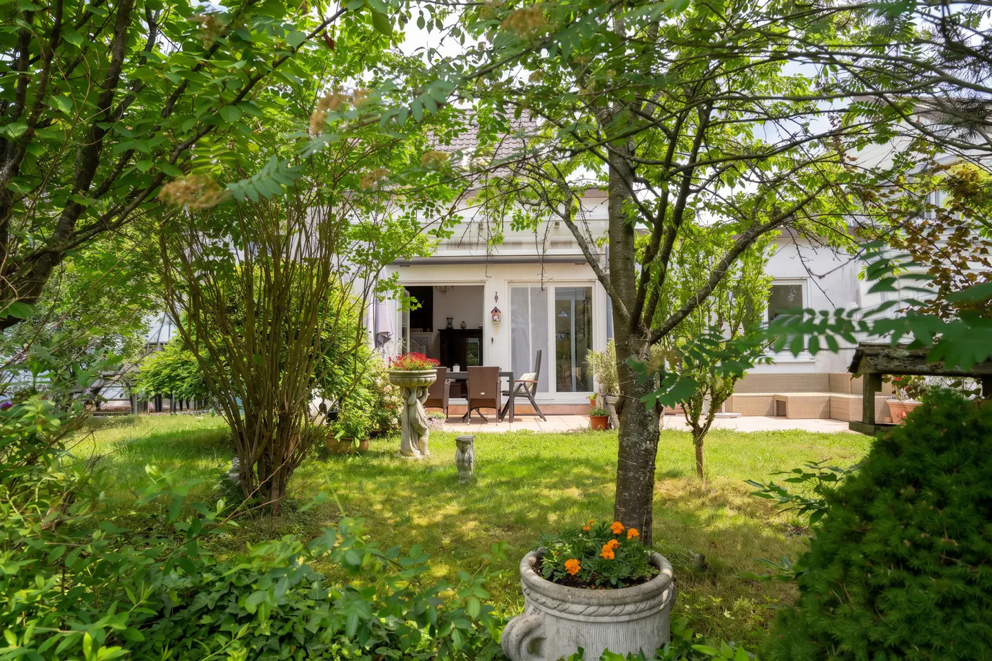 A lush green garden with a white house in the background. A patio set is visible through the open sliding doors. Flowers add pops of color.