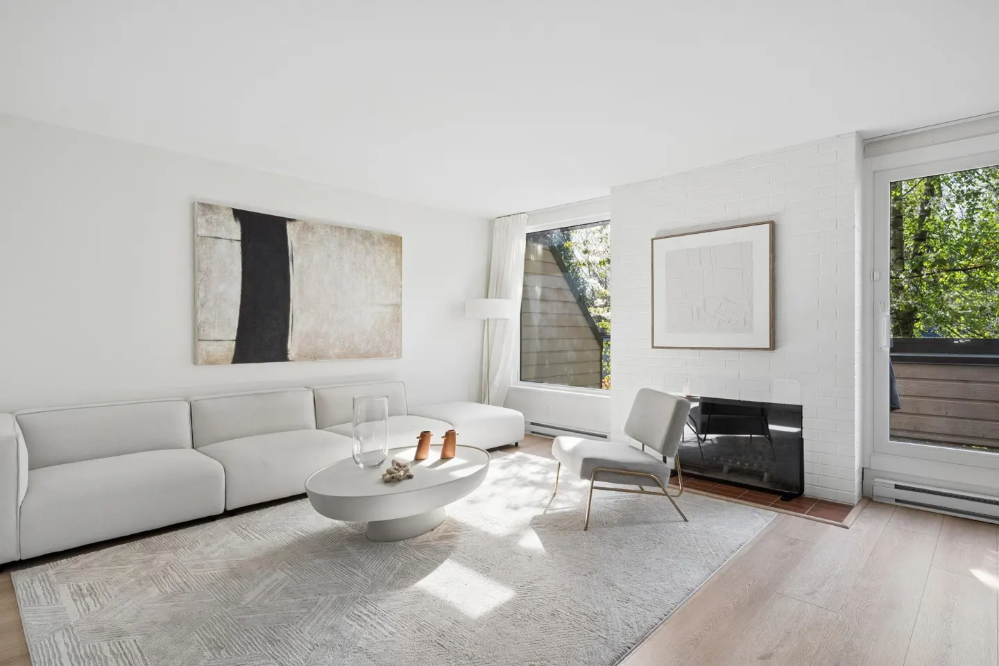 Bright living room with white walls, a gray sofa, and a round coffee table on a patterned rug. A modern chair sits near a fireplace and a window with green trees outside.
