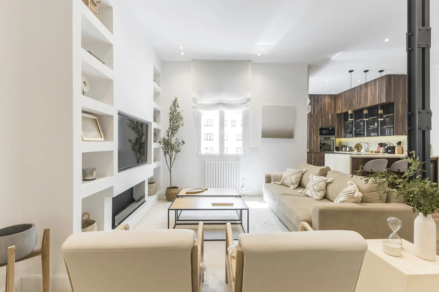 Bright living room with white built-in shelves, a TV, and a beige sofa. Two chairs face the camera. A kitchen is visible in the background.