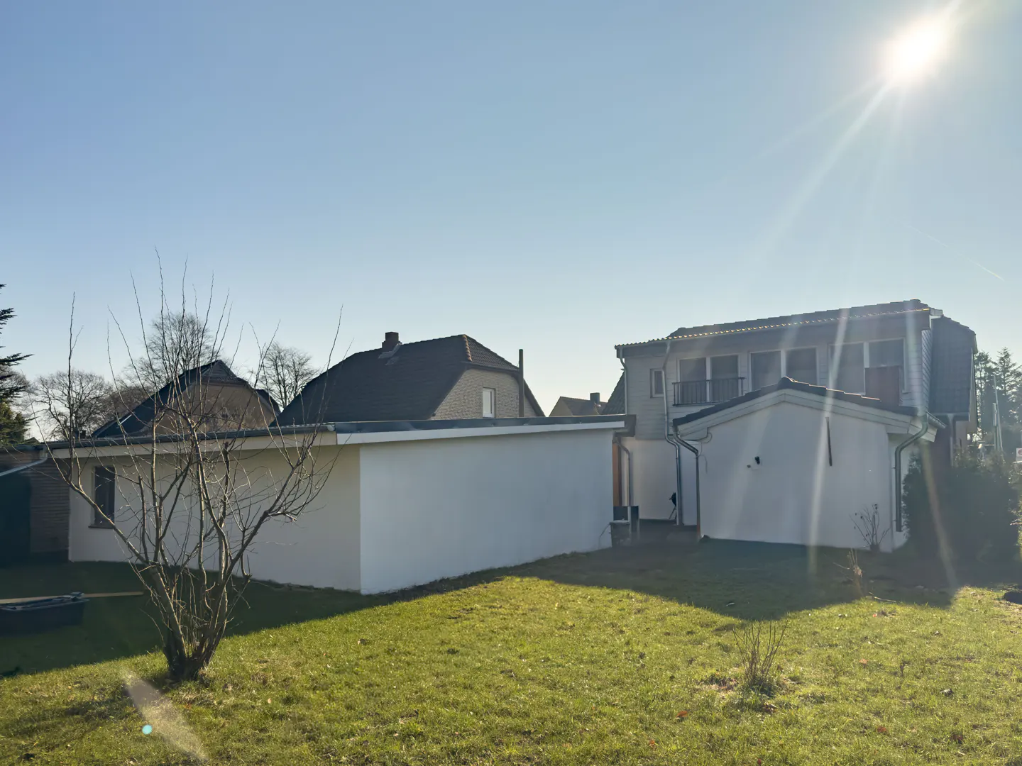 Backyard view of a two-story house with a white detached garage on a sunny day. Green grass and a bare tree are in the foreground.