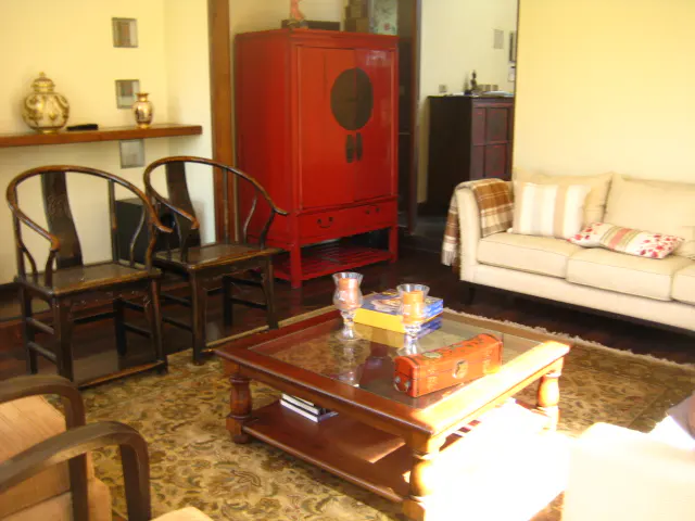 Living room with a cream sofa, wooden chairs, and a red cabinet. A glass-topped coffee table sits on a patterned rug.