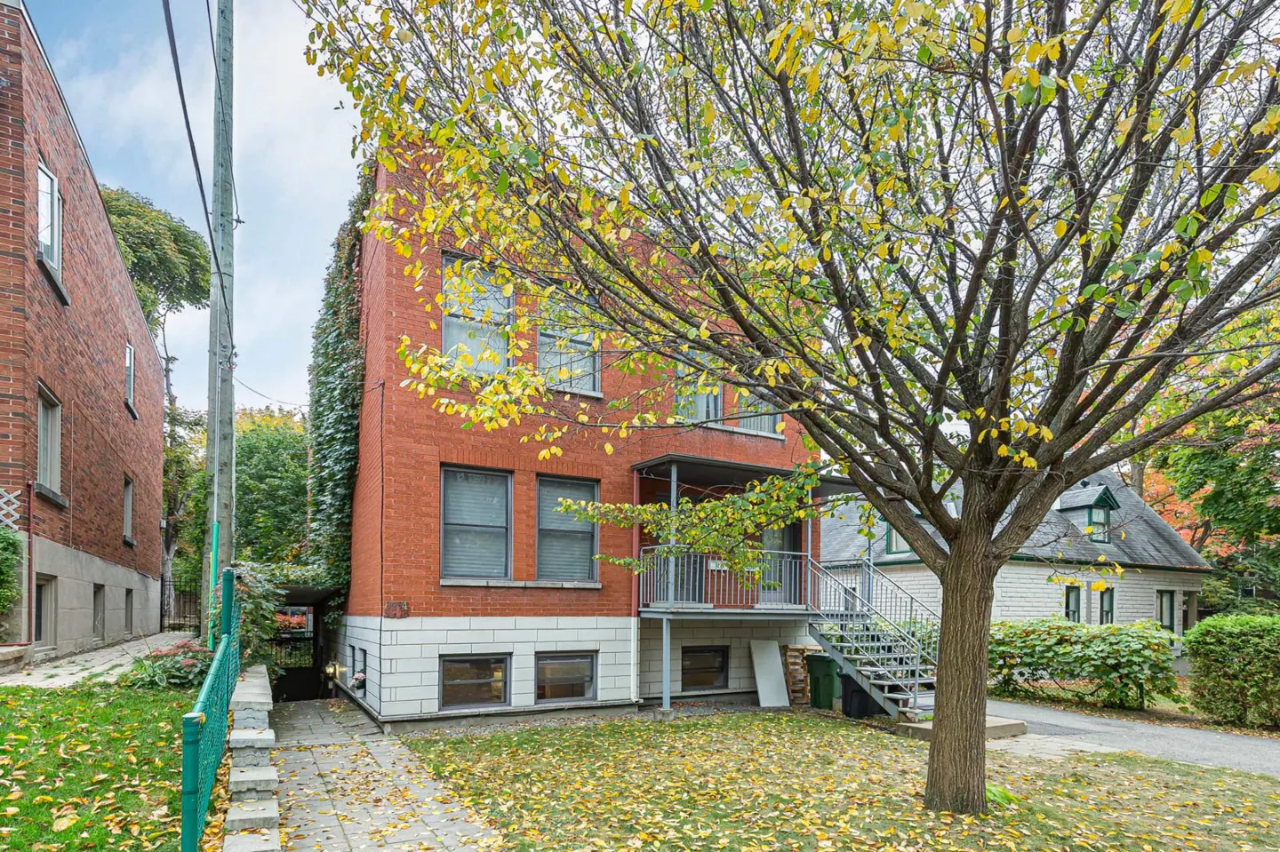 Brick apartment building with a metal staircase and balcony, framed by a tree with yellow leaves.
