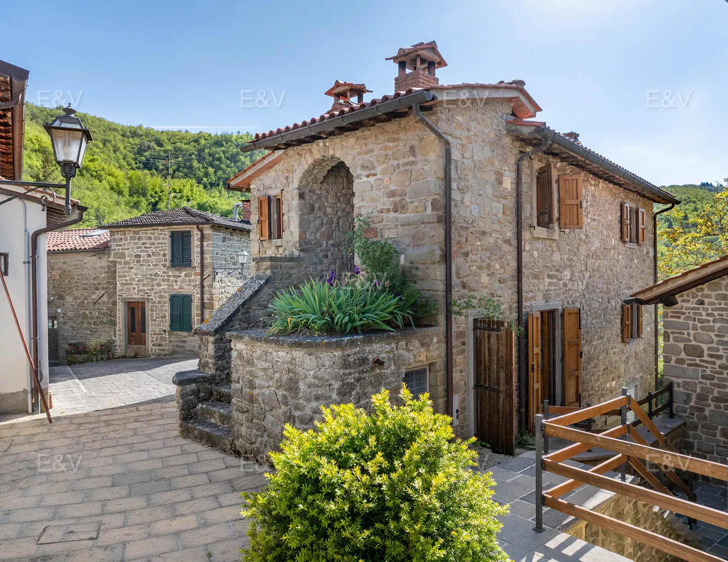 Exterior view of a stone house with brown shutters and a red tile roof in a village setting.