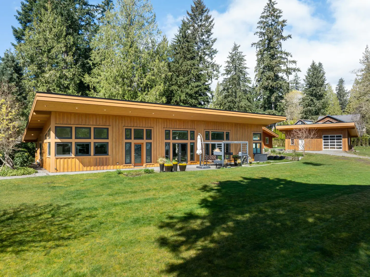 A modern, single-story wood home with large windows and a green lawn. Tall trees are in the background.