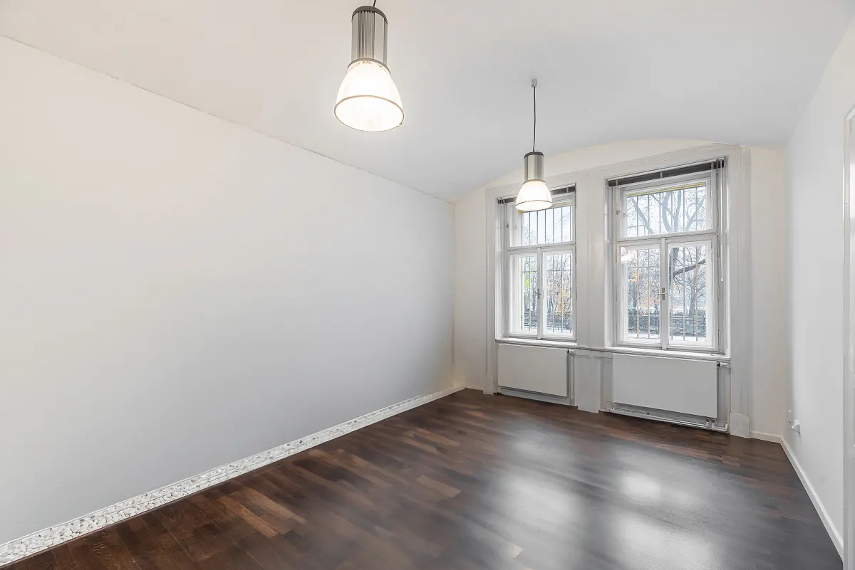 Bright, empty room with dark wood floors, white walls, and two windows. Two pendant lights hang from the ceiling.