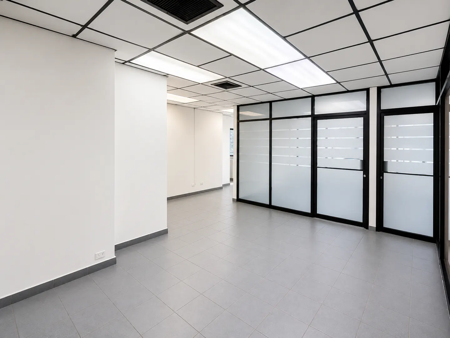 Empty modern office space with white walls, grey tiled floors, drop ceiling, and black-framed frosted glass partitions.