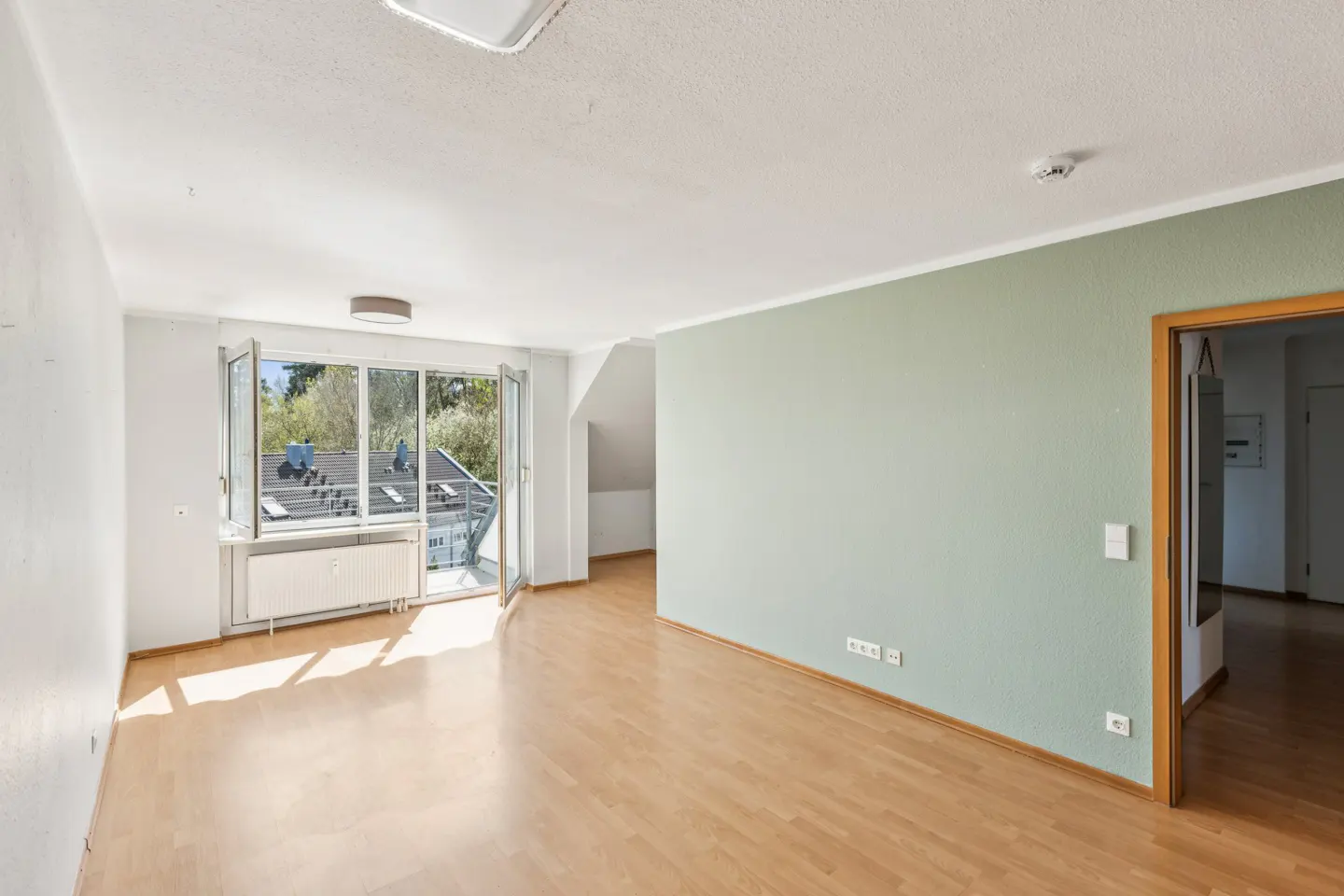 Bright, empty room with wood floors, white walls, and a light green accent wall. Balcony with open glass doors lets in sunlight.