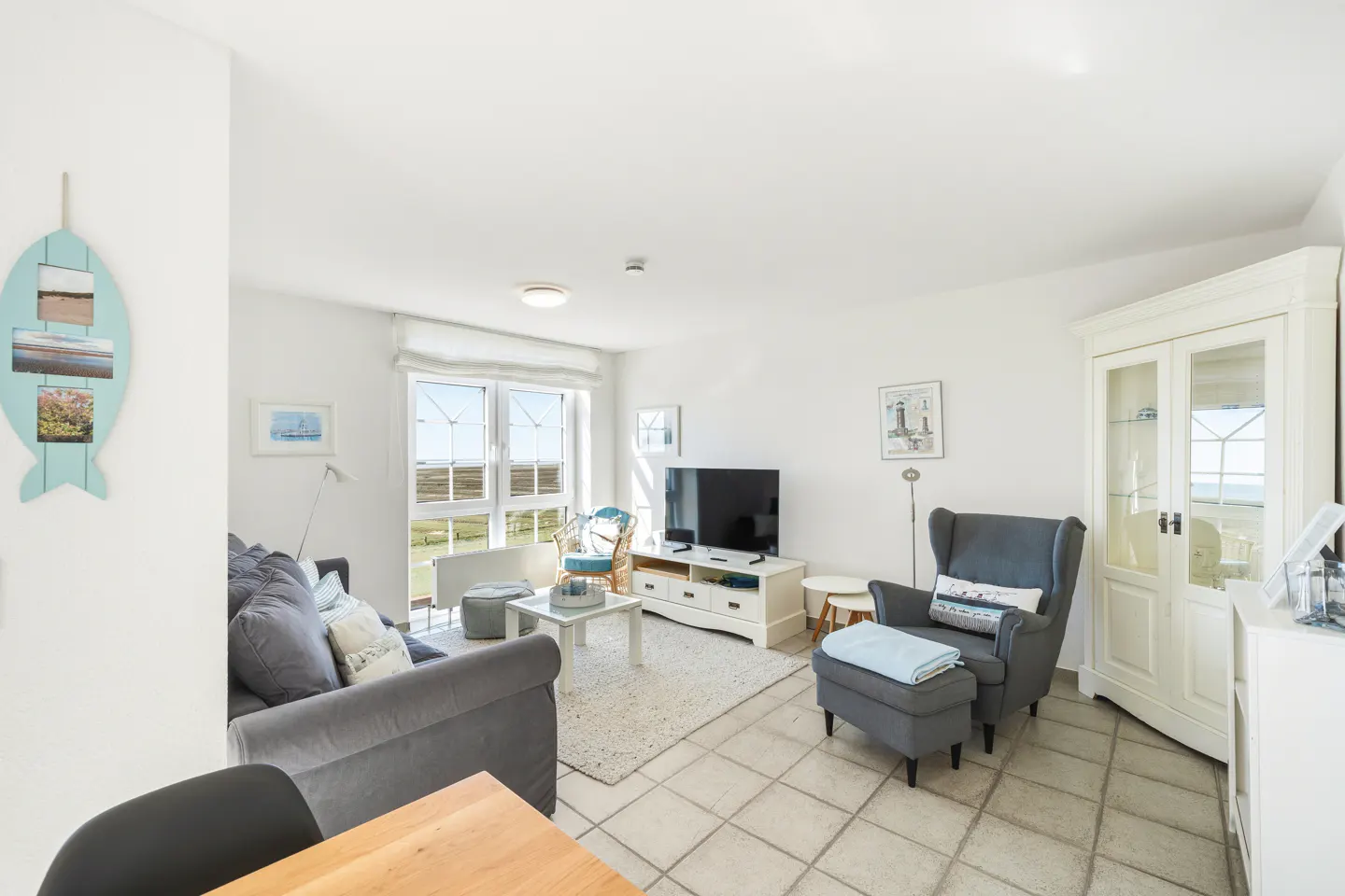 Bright living room with gray sofa, armchair, TV, and white furniture. A blue fish decoration hangs on the wall. Window view of green fields.