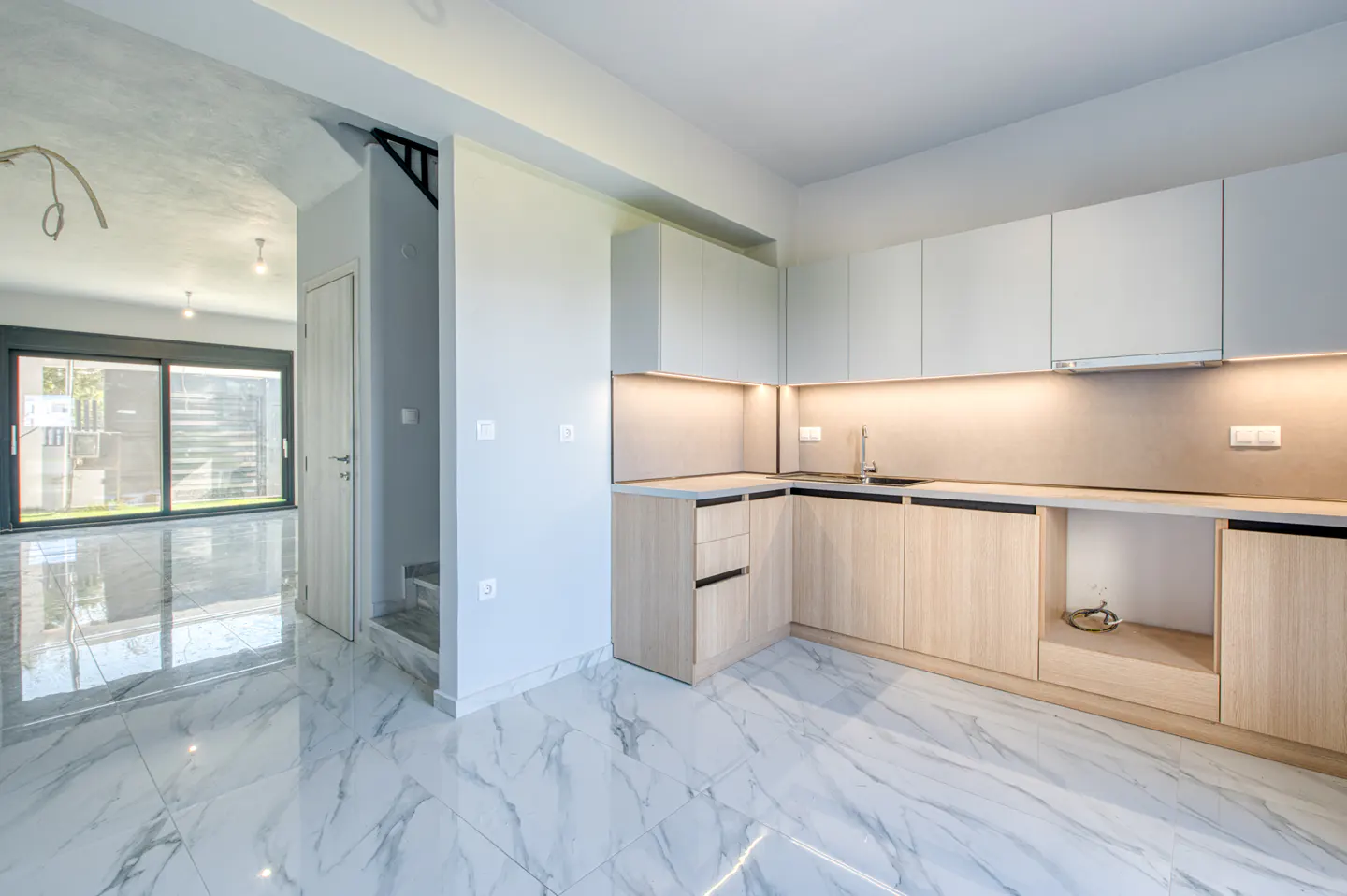 A modern kitchen with white cabinets, light wood lower cabinets, and marble floors. A large window is visible in the background.