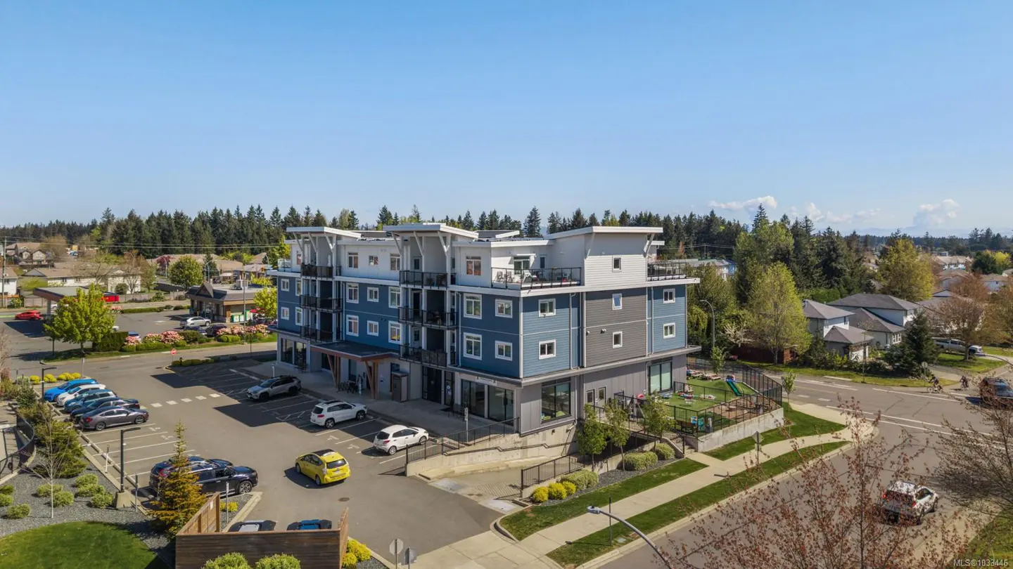 Aerial view of a modern, four-story apartment building with blue and gray siding, a parking lot, and a playground.