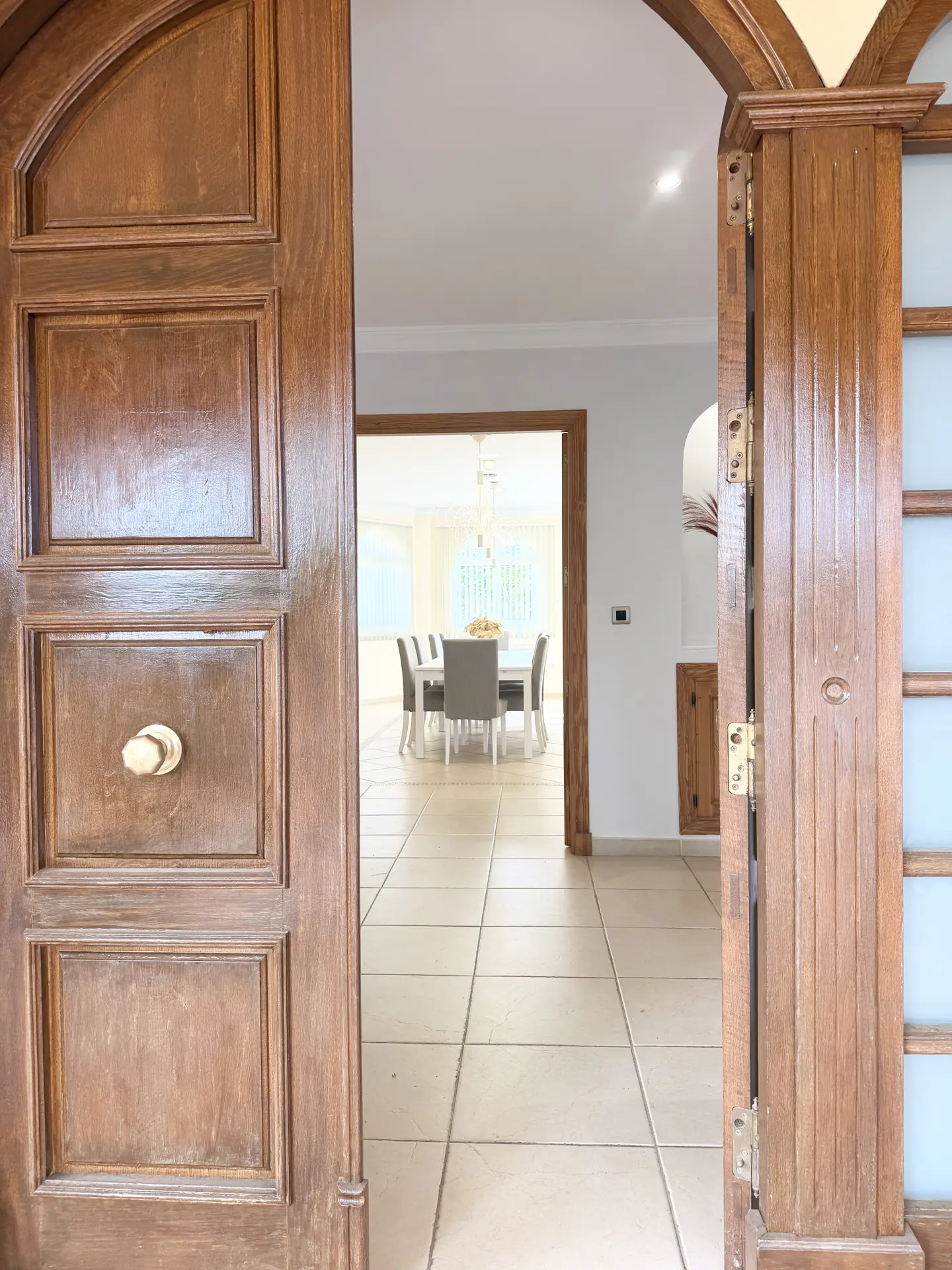 Open, carved wood double doors reveal a bright dining room with a white table and gray chairs. Tiled floor leads to the room.
