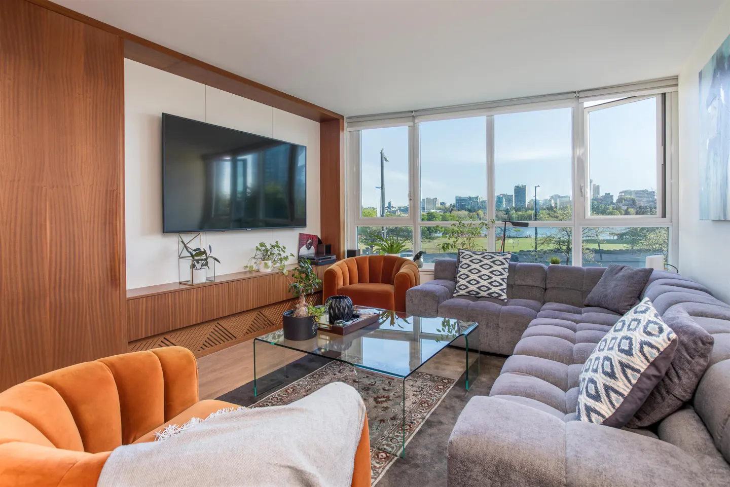 Bright living room with a large window view of a city skyline, a gray sectional sofa, and orange armchairs.