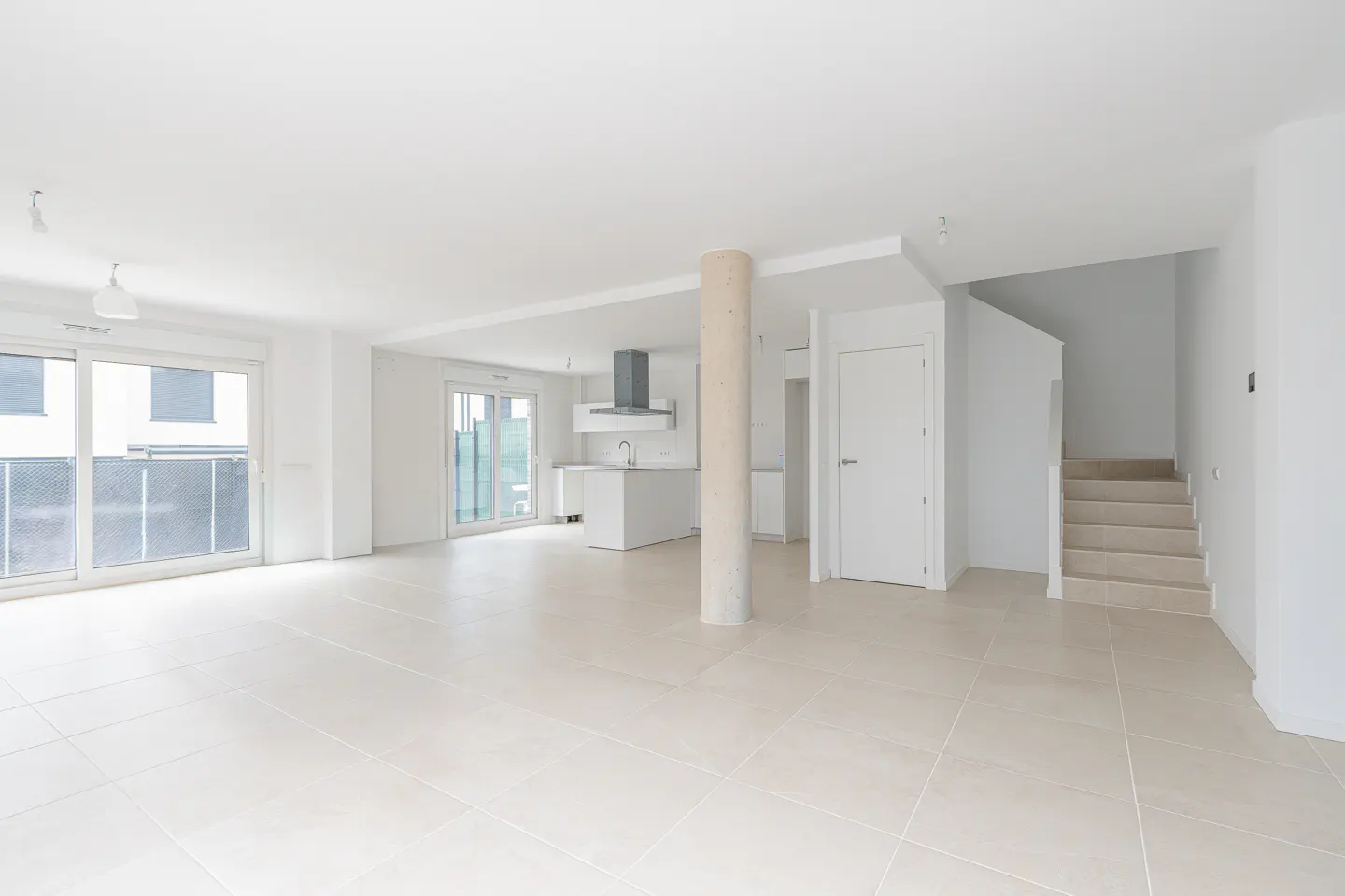 Bright, empty room with white walls, tile floor, and kitchen island. Sliding glass doors lead to a balcony. A concrete pillar stands near a white door and stairs.